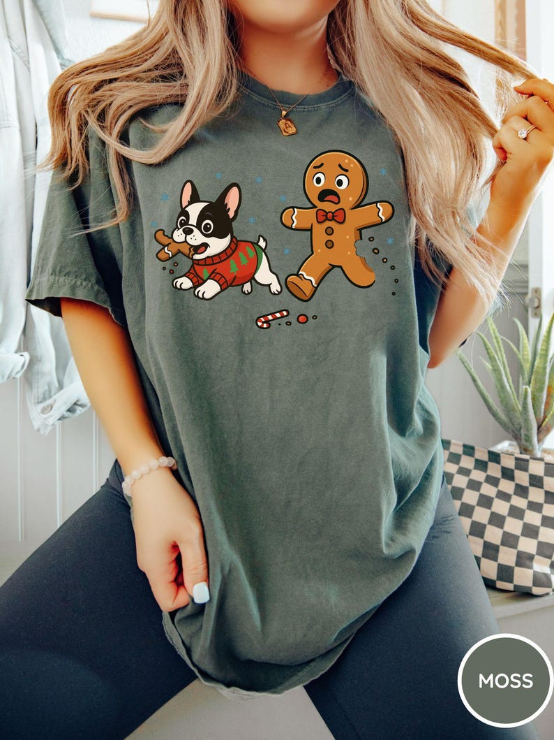 May include: A moss green t-shirt featuring a cartoon illustration of a French bulldog wearing a Christmas sweater, holding a bone, and a scared gingerbread man. The shirt has a distressed look and a round neckline.