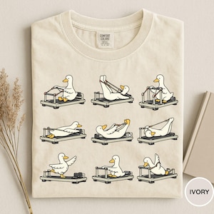 K&ouml;nnte beinhalten: Elfenbeinfarbenes T-Shirt mit Cartoon-Enten, die auf Pilates-Reformer-Ger&auml;ten trainieren. Die Enten befinden sich in verschiedenen Posen. Der Text "COMFORT COLORS" ist oben in der Mitte des Shirts zu sehen.