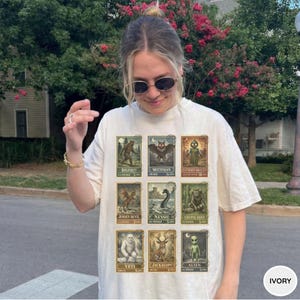 May include: Ivory-colored t-shirt featuring a collection of vintage-style postage stamps with cryptid illustrations. The stamps depict creatures like Bigfoot, Mothman, and the Jersey Devil. The shirt has a relaxed fit and short sleeves.