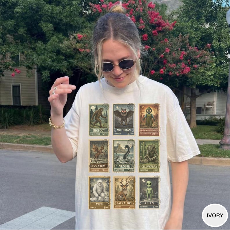 May include: Ivory-colored t-shirt featuring a collection of vintage-style postage stamps with cryptid illustrations. The stamps depict creatures like Bigfoot, Mothman, and the Jersey Devil. The shirt has a relaxed fit and short sleeves.