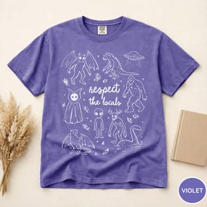 May include: Violet t-shirt with a white graphic of various cryptids and the text "respect the locals". The design includes a bat-like creature, a dinosaur, a UFO, a bigfoot, an alien, and the Loch Ness Monster.
