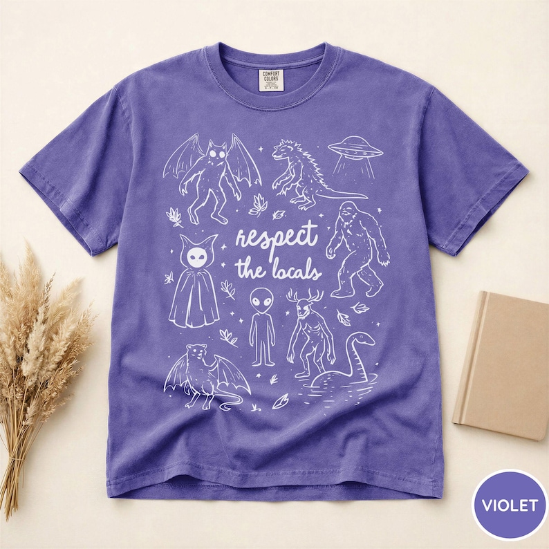 May include: Violet t-shirt with a white graphic of various cryptids and the text "respect the locals". The design includes a bat-like creature, a dinosaur, a UFO, a bigfoot, an alien, and the Loch Ness Monster.