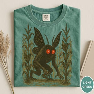 K&ouml;nnte beinhalten: Hellgr&uuml;nes T-Shirt mit einem dunklen Kreaturen-Design, roten Augen und Fl&uuml;geln, umgeben von Maisstauden. Die Grafik ist auf einem Comfort Colors-Shirt gedruckt. Das gefaltete Shirt wird auf hellem Hintergrund pr&auml;sentiert.