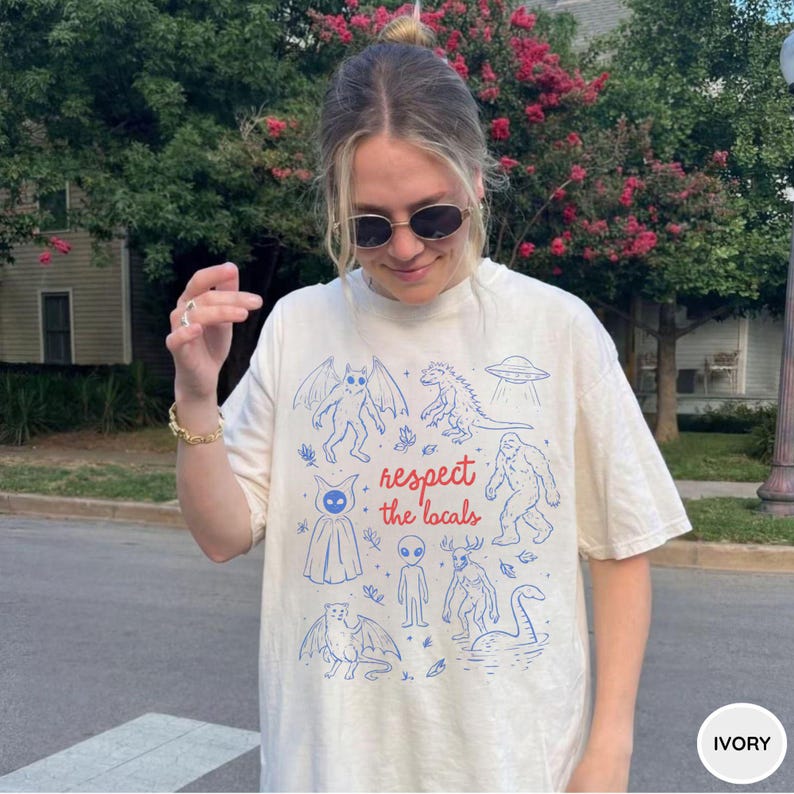 May include: Ivory-colored t-shirt featuring blue illustrations of mythical creatures and an alien, with the text "respect the locals" in red. The shirt is a relaxed fit.
