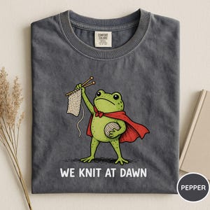 May include: Dark grey t-shirt featuring a cartoon frog superhero with a red cape, holding knitting needles and yarn. The text "WE KNIT AT DAWN" is printed below the frog. The shirt is made of a soft material.