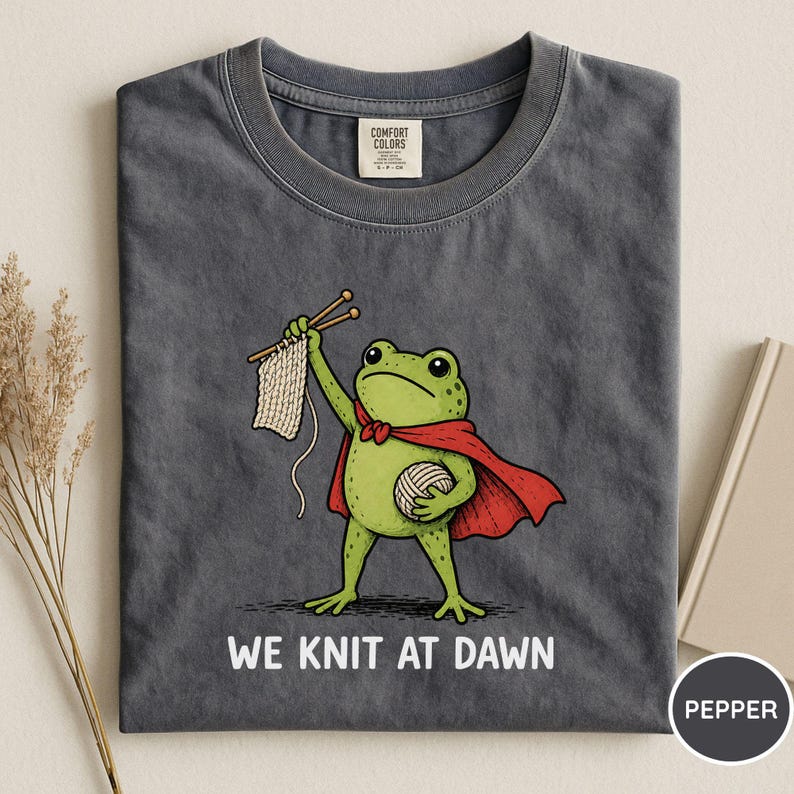 May include: Dark grey t-shirt featuring a cartoon frog superhero with a red cape, holding knitting needles and yarn. The text "WE KNIT AT DAWN" is printed below the frog. The shirt is made of a soft material.