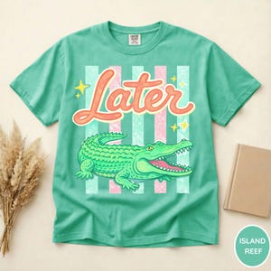 May include: A teal t-shirt with a graphic of a green alligator and the word "Later" in orange script. The design includes pink and teal stripes and yellow stars. The shirt is a Comfort Colors brand.