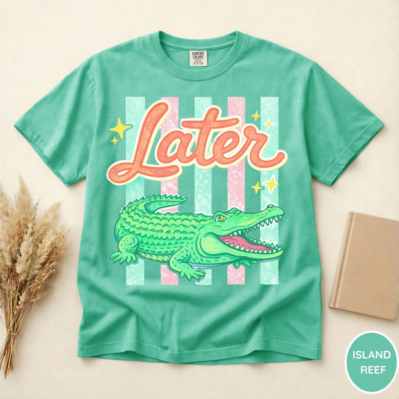 May include: A teal t-shirt with a graphic of a green alligator and the word "Later" in orange script. The design includes pink and teal stripes and yellow stars. The shirt is a Comfort Colors brand.