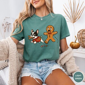 May include: Light green t-shirt featuring a cartoon dog in a Christmas sweater holding a gingerbread man. A gingerbread man is running away, with a candy cane on the ground. The shirt is a light green color.