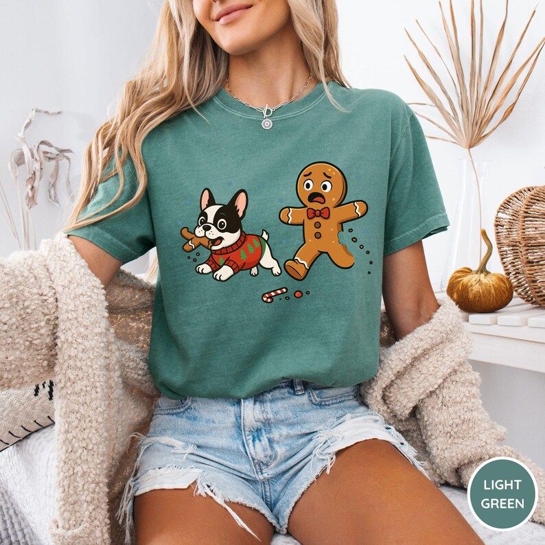 May include: Light green t-shirt featuring a cartoon dog in a Christmas sweater holding a gingerbread man. A gingerbread man is running away, with a candy cane on the ground. The shirt is a light green color.