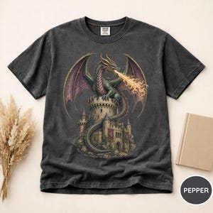 May include: A charcoal gray short-sleeve t-shirt featuring a graphic print of a green dragon perched atop a stone castle tower, breathing a stream of orange fire. The dragon has large purple wings and a long tail wrapped around the castle structure.