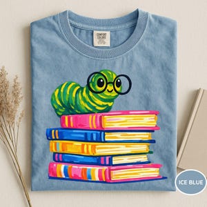 Pu&ograve; includere: Una t-shirt color blu ghiaccio a girocollo con la grafica di un bruco verde che indossa occhiali tondi neri, appoggiato sopra una pila di cinque libri colorati. La maglietta presenta un'etichetta del marchio Comfort Colors all'interno del colletto.