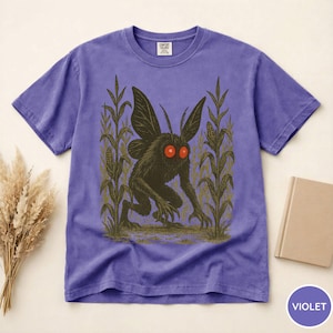 K&ouml;nnte beinhalten: Violettes T-Shirt mit einem Motiv einer dunklen Kreatur mit roten Augen und gro&szlig;en Fl&uuml;geln, vor dem Hintergrund von Maispflanzen. Das T-Shirt hat einen Rundhalsausschnitt und kurze &Auml;rmel. Das Shirt ist aus weichem Material.
