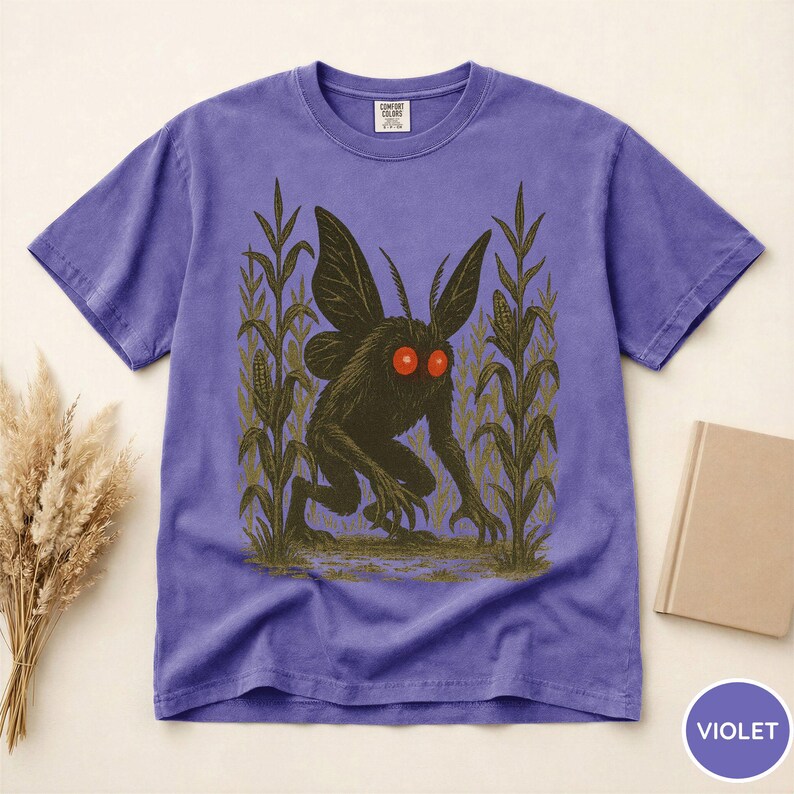 K&ouml;nnte beinhalten: Violettes T-Shirt mit einem Motiv einer dunklen Kreatur mit roten Augen und gro&szlig;en Fl&uuml;geln, vor dem Hintergrund von Maispflanzen. Das T-Shirt hat einen Rundhalsausschnitt und kurze &Auml;rmel. Das Shirt ist aus weichem Material.