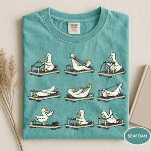 K&ouml;nnte beinhalten: T&uuml;rkisfarbenes T-Shirt mit einem sich wiederholenden Muster von Cartoon-Enten, die auf Pilates-Reformern trainieren. Die Enten sind wei&szlig; mit gelben Schn&auml;beln und F&uuml;&szlig;en. Das Shirt hat einen Rundhalsausschnitt und ein Comfort Colors-Etikett.