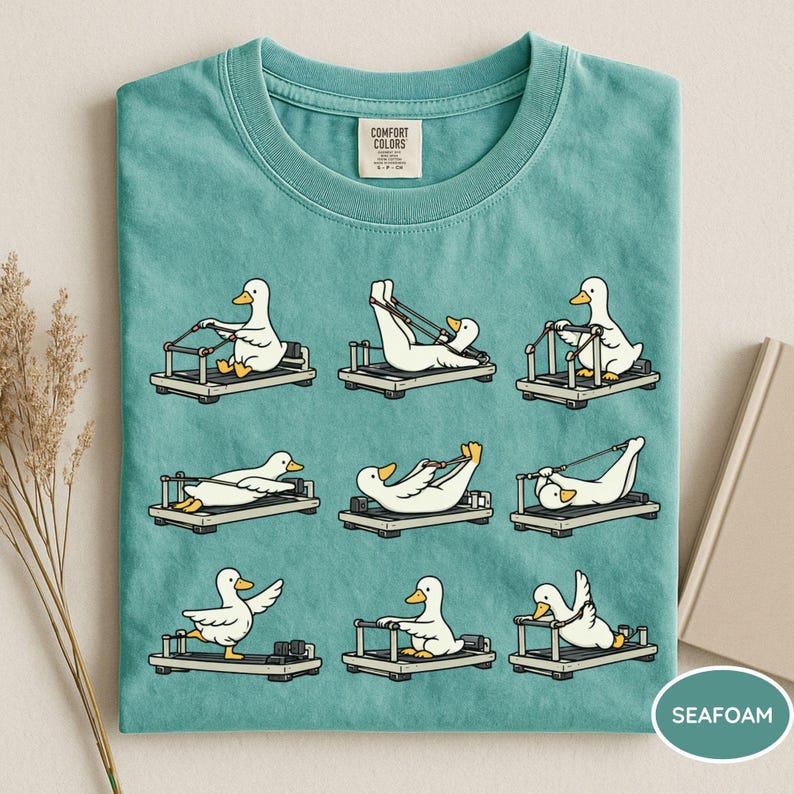K&ouml;nnte beinhalten: T&uuml;rkisfarbenes T-Shirt mit einem sich wiederholenden Muster von Cartoon-Enten, die auf Pilates-Reformern trainieren. Die Enten sind wei&szlig; mit gelben Schn&auml;beln und F&uuml;&szlig;en. Das Shirt hat einen Rundhalsausschnitt und ein Comfort Colors-Etikett.