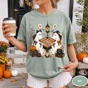 May include: A sage green t-shirt featuring a graphic of four ghosts playing mahjong around a table. The design includes a hanging lantern, a pumpkin, stacks of books, and mahjong tiles. The ghosts wear bows and hats, surrounded by floral accents.