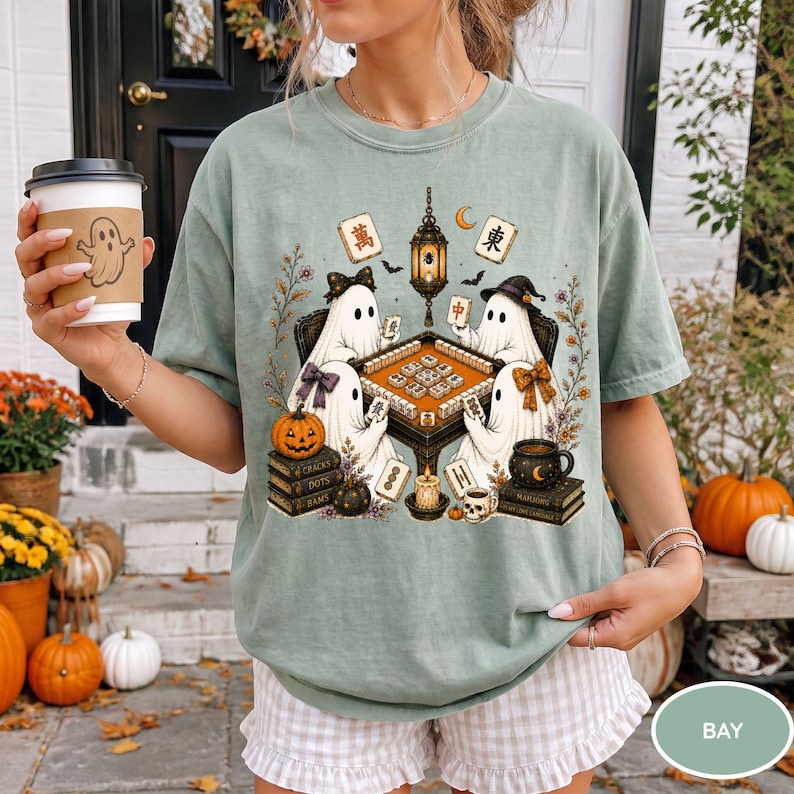 May include: A sage green t-shirt featuring a graphic of four ghosts playing mahjong around a table. The design includes a hanging lantern, a pumpkin, stacks of books, and mahjong tiles. The ghosts wear bows and hats, surrounded by floral accents.