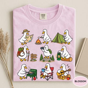 May include: A light pink t-shirt featuring a graphic print of nine white ducks engaged in various camping activities, including reading a map, roasting marshmallows, setting up a tent, hiking, using binoculars, holding a lantern, cooking, and playing guitar.