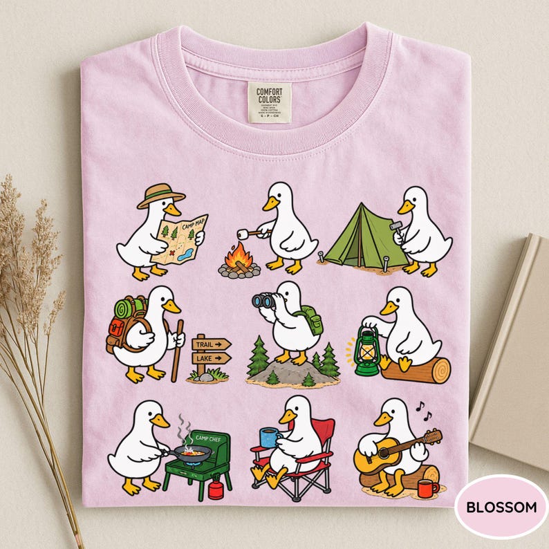 May include: A light pink t-shirt featuring a graphic print of nine white ducks engaged in various camping activities, including reading a map, roasting marshmallows, setting up a tent, hiking, using binoculars, holding a lantern, cooking, and playing guitar.
