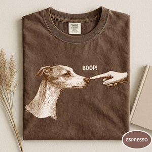 May include: A brown t-shirt featuring a graphic illustration of a dog being touched on the nose by a human finger. The text BOOP! is printed above the dog's nose. The shirt has a Comfort Colors label at the inner neckline and a solid brown color.