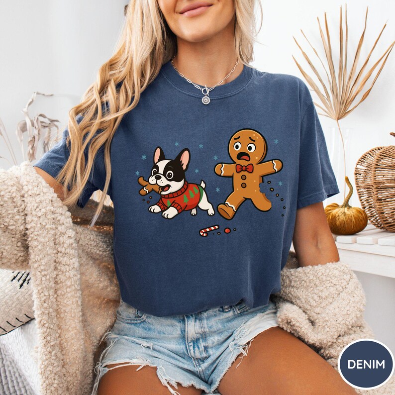May include: Denim blue t-shirt featuring a cartoon dog in a Christmas sweater with a bone and a gingerbread man with a worried expression. The design includes snowflakes and a broken candy cane.