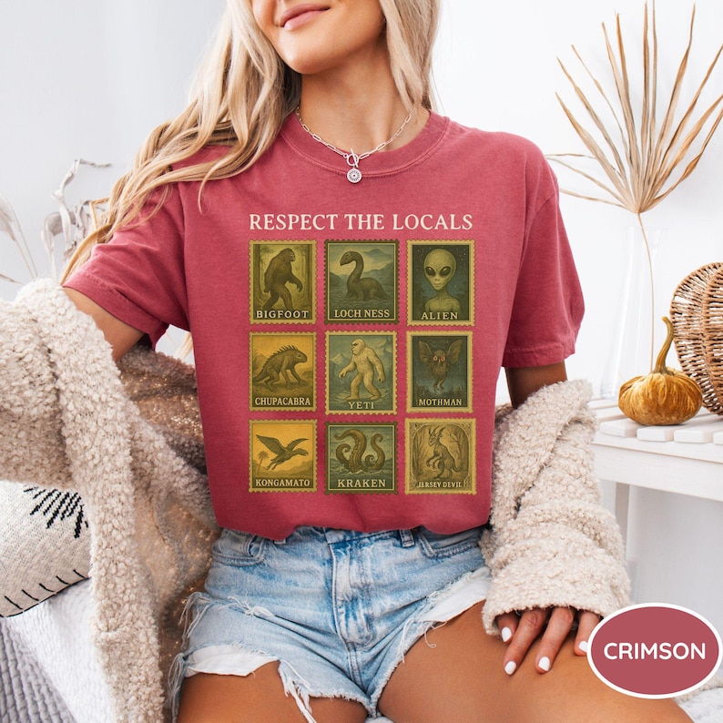 Pu&ograve; includere: T-shirt color cremisi con la scritta "RESPECT THE LOCALS" e illustrazioni di criptidi come Bigfoot e il mostro di Loch Ness in un design a francobollo. La maglietta &egrave; realizzata in un materiale morbido.