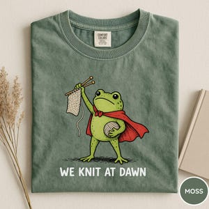 May include: Moss green t-shirt featuring a cartoon frog superhero with a red cape, holding knitting needles and yarn. The text "WE KNIT AT DAWN" is printed below the frog. The shirt is folded and has a Comfort Colors tag.