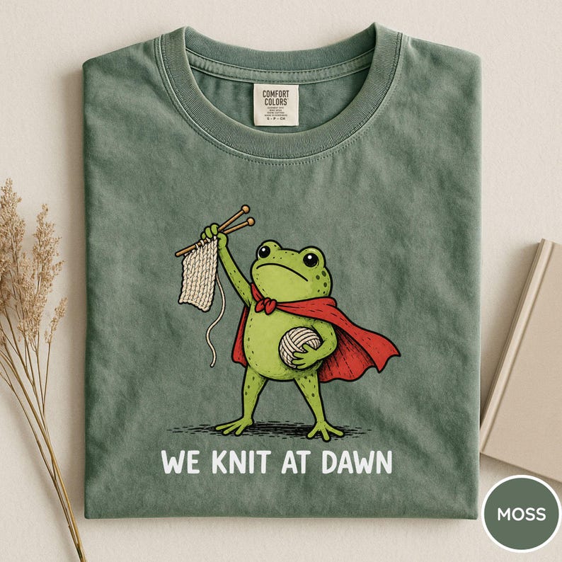 May include: Moss green t-shirt featuring a cartoon frog superhero with a red cape, holding knitting needles and yarn. The text "WE KNIT AT DAWN" is printed below the frog. The shirt is folded and has a Comfort Colors tag.