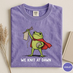 May include: Violet Comfort Colors t-shirt featuring a cartoon frog superhero with a red cape, holding knitting needles and yarn. The text "WE KNIT AT DAWN" is printed below the frog. A book and dried plant are in the background.