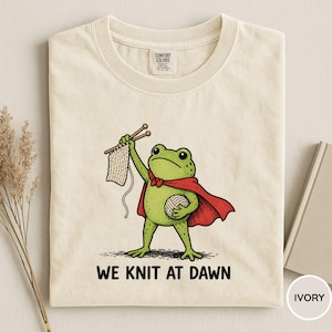May include: Ivory-colored t-shirt featuring a cartoon frog in a red cape, holding knitting needles and yarn. The frog is knitting a piece of fabric. The text "WE KNIT AT DAWN" is printed below the frog.