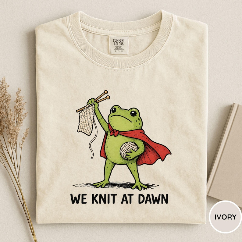 May include: Ivory-colored t-shirt featuring a cartoon frog in a red cape, holding knitting needles and yarn. The frog is knitting a piece of fabric. The text "WE KNIT AT DAWN" is printed below the frog.
