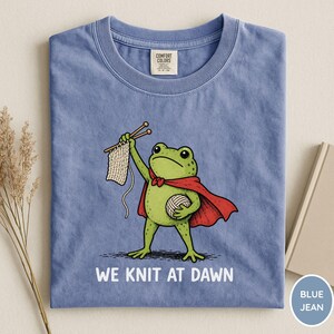 May include: Blue jean t-shirt featuring a cartoon frog superhero with a red cape, holding knitting needles and yarn. The text "WE KNIT AT DAWN" is printed below the frog. The shirt has a crew neck and a Comfort Colors tag.