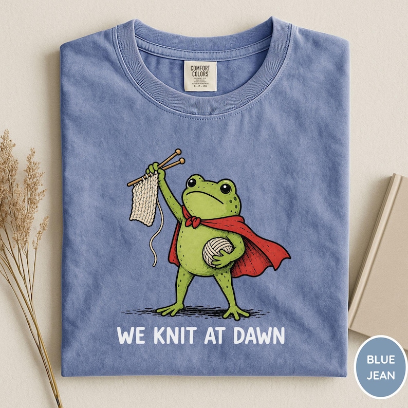 May include: Blue jean t-shirt featuring a cartoon frog superhero with a red cape, holding knitting needles and yarn. The text "WE KNIT AT DAWN" is printed below the frog. The shirt has a crew neck and a Comfort Colors tag.