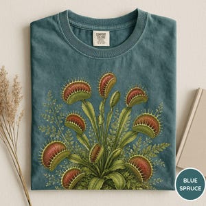 May include: Blue spruce t-shirt with a detailed illustration of Venus flytraps. The graphic shows the plant's red-edged traps and green leaves, set against ferns. The shirt has a crew neck and a tag that says "Comfort Colors".