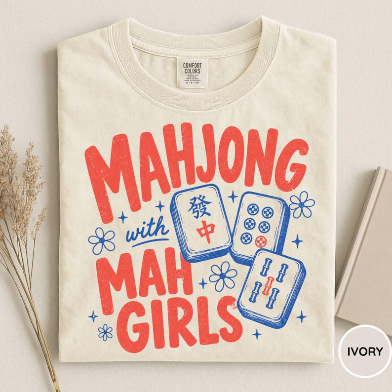 May include: An ivory t-shirt featuring the red text "MAHJONG with MAH GIRLS" and a graphic of three blue and white mahjong tiles. The design includes small blue flower and star accents. The shirt has a Comfort Colors brand label at the neck.