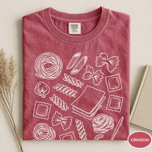 Puede incluir: Una camiseta de manga corta color carmes&iacute; con un estampado gr&aacute;fico de l&iacute;neas blancas de varias formas de pasta, incluyendo penne, farfalle, raviolis y l&aacute;minas de lasa&ntilde;a. La etiqueta interior del cuello muestra la marca Comfort Colors.