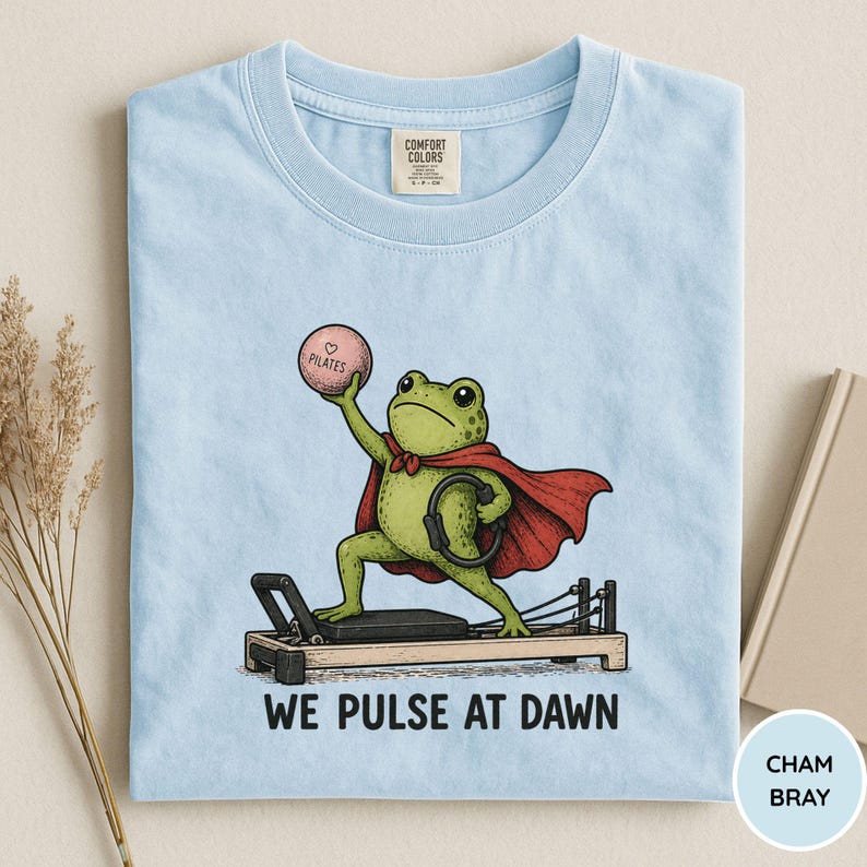 May include: A light blue t-shirt featuring a graphic of a green frog wearing a red cape, standing on a Pilates reformer machine. The frog holds a pink ball labeled Pilates and a black ring. The text below reads WE PULSE AT DAWN in black letters.