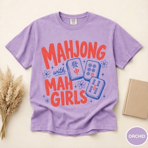 May include: A light purple short-sleeve t-shirt featuring the red and blue text "MAHJONG with MAH GIRLS" alongside illustrations of three mahjong tiles and small flower icons. The shirt has a crew neckline and a standard fit design.