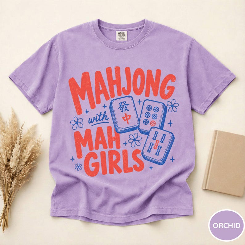 May include: A light purple short-sleeve t-shirt featuring the red and blue text "MAHJONG with MAH GIRLS" alongside illustrations of three mahjong tiles and small flower icons. The shirt has a crew neckline and a standard fit design.