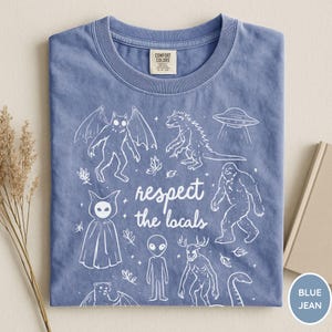 May include: Blue jean colored t-shirt with a crew neck. The shirt features a white graphic of various cryptids and a UFO, with the text "respect the locals". The shirt is made of a soft material.