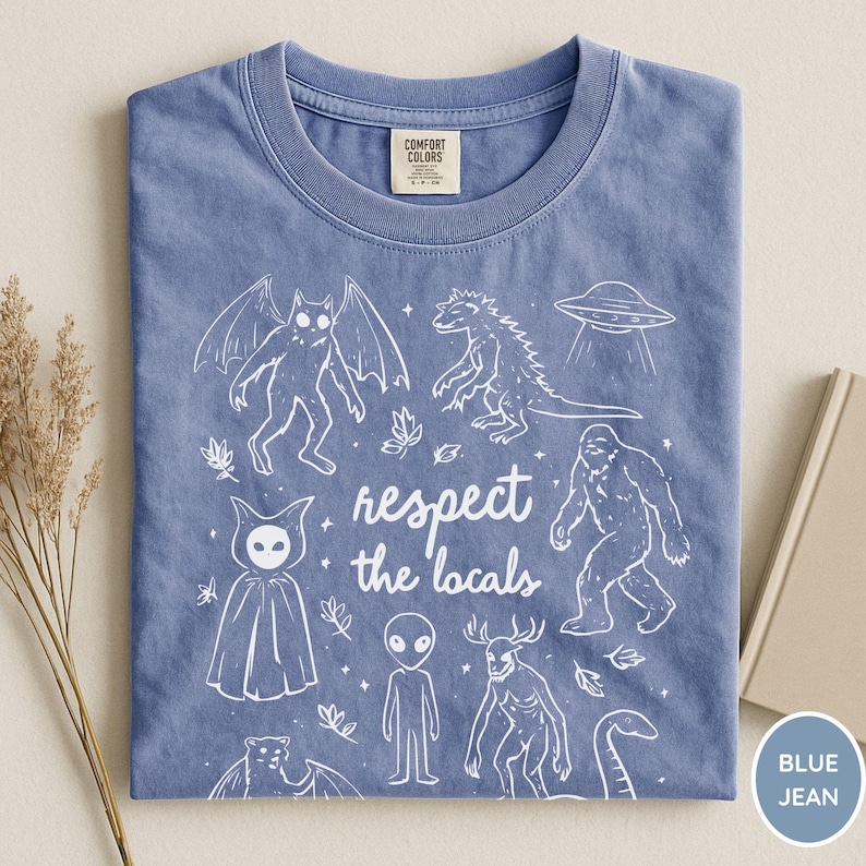 May include: Blue jean colored t-shirt with a crew neck. The shirt features a white graphic of various cryptids and a UFO, with the text "respect the locals". The shirt is made of a soft material.