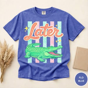 May include: A blue t-shirt with a graphic of a green alligator and the word "Later" in orange script. The design includes vertical stripes in light blue and pink, with yellow star accents. The shirt is a Comfort Colors brand.
