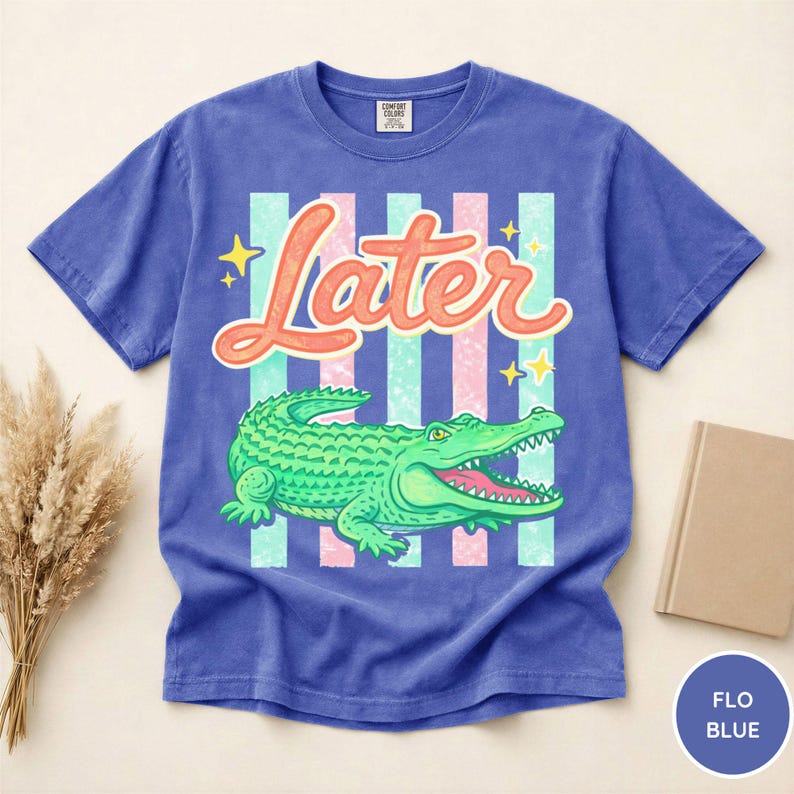 May include: A blue t-shirt with a graphic of a green alligator and the word "Later" in orange script. The design includes vertical stripes in light blue and pink, with yellow star accents. The shirt is a Comfort Colors brand.