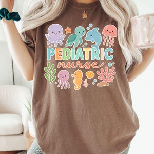 K&ouml;nnte beinhalten: Espressofarbenes T-Shirt mit einem farbenfrohen Aufdruck mit Meerestieren und dem Schriftzug "Pediatric Nurse". Das Design umfasst einen Oktopus, eine Schildkr&ouml;te, einen Delfin, eine Qualle, ein Seepferdchen und Korallen. Das Shirt ist ein l&auml;ssiger, bequemer Stil.