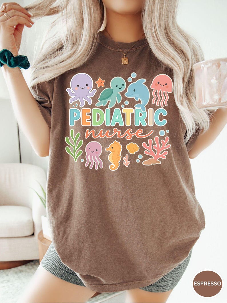 K&ouml;nnte beinhalten: Espressofarbenes T-Shirt mit einem farbenfrohen Aufdruck mit Meerestieren und dem Schriftzug "Pediatric Nurse". Das Design umfasst einen Oktopus, eine Schildkr&ouml;te, einen Delfin, eine Qualle, ein Seepferdchen und Korallen. Das Shirt ist ein l&auml;ssiger, bequemer Stil.