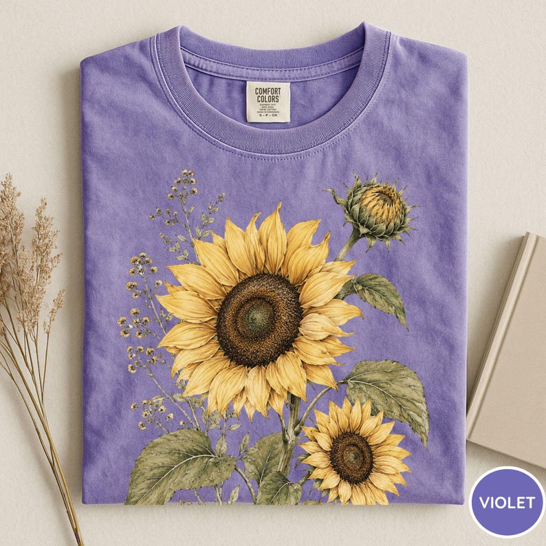 May include: A violet Comfort Colors t-shirt featuring a detailed watercolor illustration of sunflowers. The design includes a large, open sunflower, a smaller one, and a closed bud, with green leaves and delicate sprigs.
