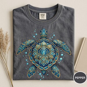 May include: A charcoal gray t-shirt featuring a central graphic of a sea turtle with an intricate blue and gold mandala-style shell pattern. The design includes small blue bubbles surrounding the turtle. The shirt has a Comfort Colors brand label.