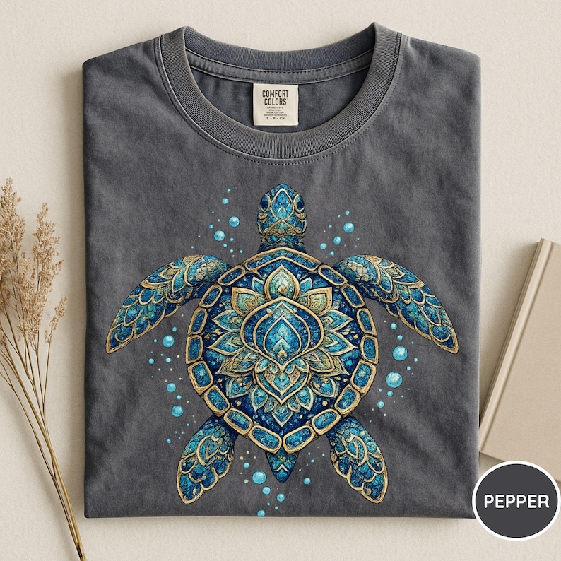 May include: A charcoal gray t-shirt featuring a central graphic of a sea turtle with an intricate blue and gold mandala-style shell pattern. The design includes small blue bubbles surrounding the turtle. The shirt has a Comfort Colors brand label.