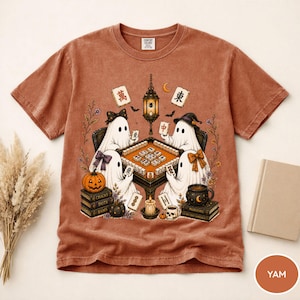 May include: A rust-colored short-sleeve t-shirt featuring a graphic of four ghosts playing mahjong around a table. The design includes a hanging lantern, a pumpkin, stacks of books, a skull, and floral elements. The ghosts wear bows and hats.